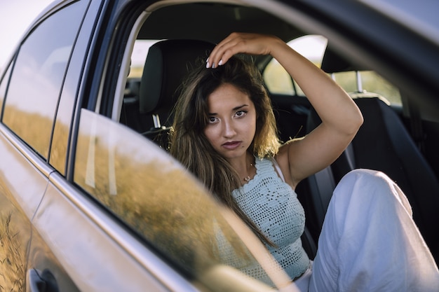 Disparo horizontal de una hermosa joven mujer caucásica posando en el asiento delantero de un coche en un campo