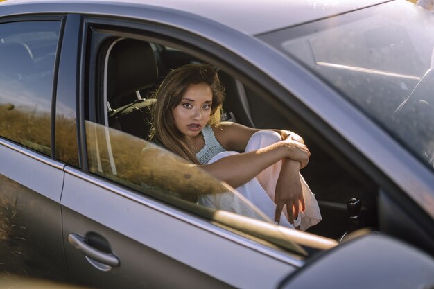 Disparo horizontal de una hermosa joven mujer caucásica posando en el asiento delantero de un coche en un campo