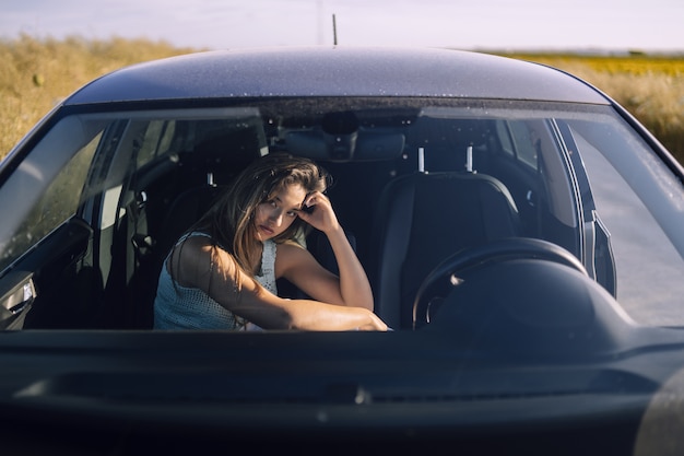 Disparo horizontal de una hermosa joven mujer caucásica posando en el asiento delantero de un coche en un campo