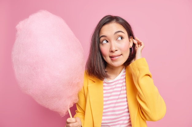 Disparo horizontal de hermosa morena asiática adolescente mira hacia otro lado con expresión pensativa sostiene delicioso algodón de azúcar viste chaqueta amarilla aislada sobre pared rosa. Concepto goloso