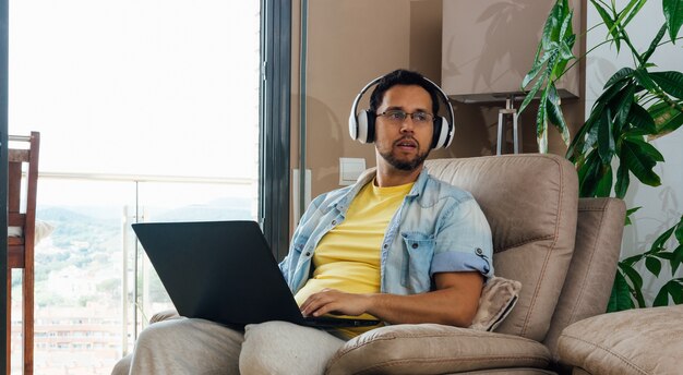 Disparo horizontal de un hombre escuchando música con auriculares y portátil de rodillas