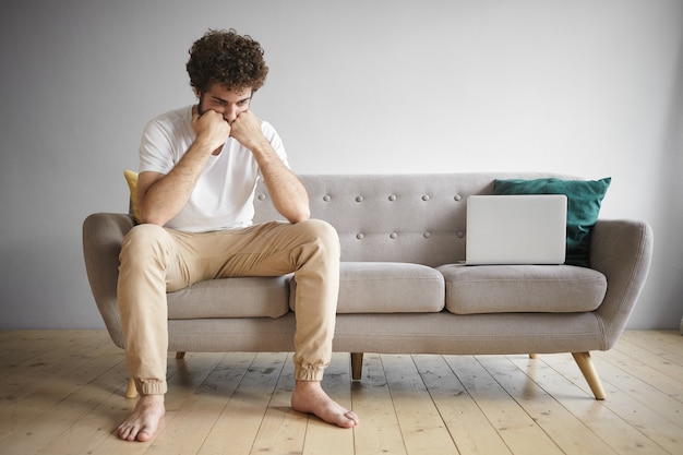 Disparo horizontal de hombre joven desempleado con camiseta blanca y jeans beige sentado descalzo en el sofá computadora portátil de trabajo con expresión facial triste y frustrada, buscando trabajo en línea