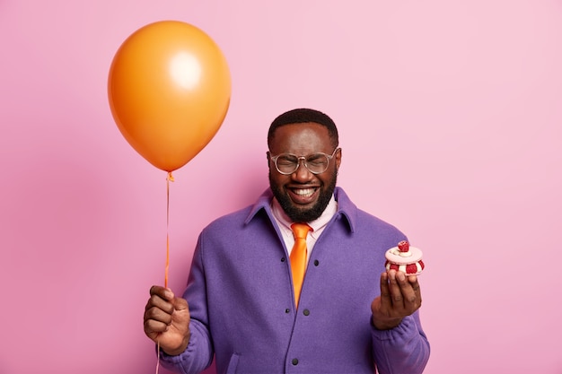 Disparo horizontal de hombre negro sostiene airballoon inflado, cupcake cremoso, se ríe sinceramente, felicita a un amigo con la graduación, tiene fiesta