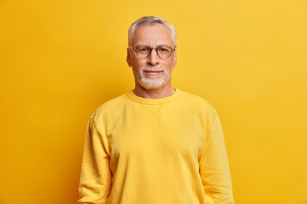 Disparo horizontal de hombre de pelo gris con arrugas lleva gafas y un jersey amarillo casual mira directamente al frente tiene poses de expresión satisfecha en el interior