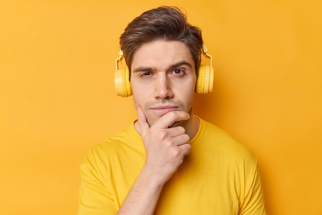 Disparo horizontal de un hombre pensativo sostiene la barbilla y mira seriamente a la cámara escucha su canción favorita a través de auriculares estéreo vestidos con ropa informal posa contra un fondo amarillo vívido Déjame pensar