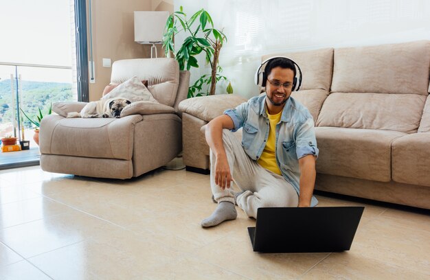 Disparo horizontal de un hombre sentado en el suelo escuchando música y trabajando con un portátil en casa