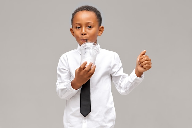 Disparo horizontal de lindo niño de piel oscura con camisa blanca y corbata negra disfrutando de jugo fresco. Apuesto alumno afroamericano bebiendo batido o batido de vidrio de plástico con paja