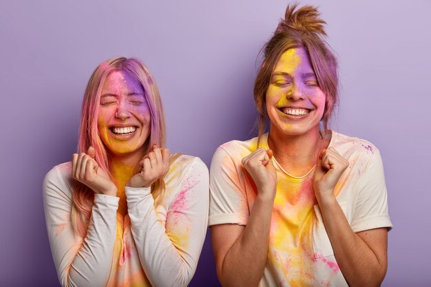 Disparo horizontal de mejores amigas llenas de alegría, apretar los puños con triunfo, celebrar el festival de Holi en la India, jugar con polvos coloridos, ropa informal manchada de blanco Celebración de la primavera que viene