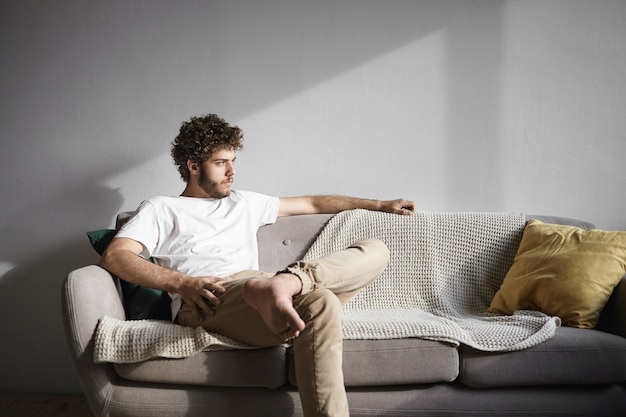 Disparo horizontal de moda joven soltero caucásico de treinta años con cabello ondulado y barba espesa sentado cómodamente en el sofá en la sala de estar, con mirada seria, tratando de relajarse después del trabajo