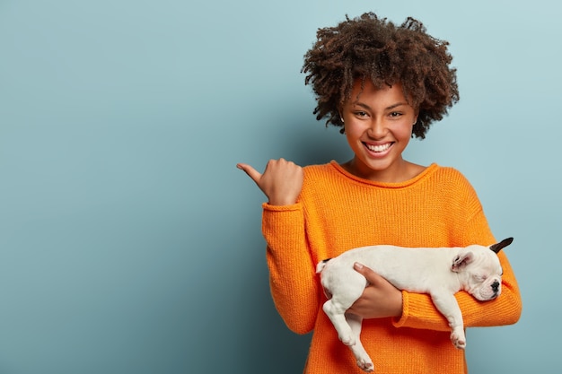 Disparo horizontal de una mujer afro complacida que señala el espacio libre, muestra la dirección a la tienda de mascotas, compró un cachorro de pedigrí, tiene una sonrisa agradable en los modelos faciales sobre una pared azul. Amor entre perro y dueño