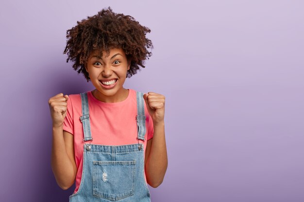 Disparo horizontal de una mujer afro negra llena de alegría que aprieta los puños con triunfo, celebra el éxito, expresa buenas emociones, se viste con ropa informal, posa sobre la pared púrpura, espera anunciar resultados