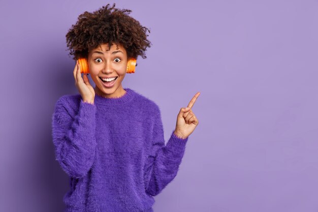 Disparo horizontal de mujer afroamericana bastante alegre escucha música agradable en los puntos de los auriculares a la derecha lleva poses de puente casual