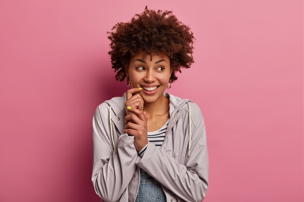 Disparo horizontal de una mujer afroamericana de pelo bastante rizado que se ve positivamente a un lado, tiene una sonrisa tierna, usa un anorak casual, mira hacia otro lado con alegría, está de buen humor, aislado en la pared rosa