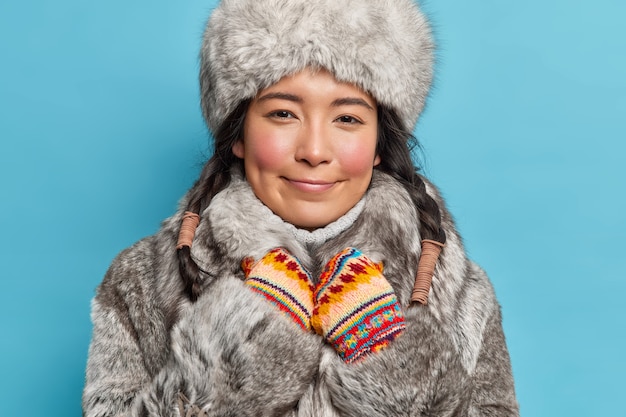 Foto gratuita disparo horizontal de mujer asiática satisfecha mira felizmente al frente viste gorro de piel y abrigo disfruta de un fantástico invierno aislado sobre una pared azul