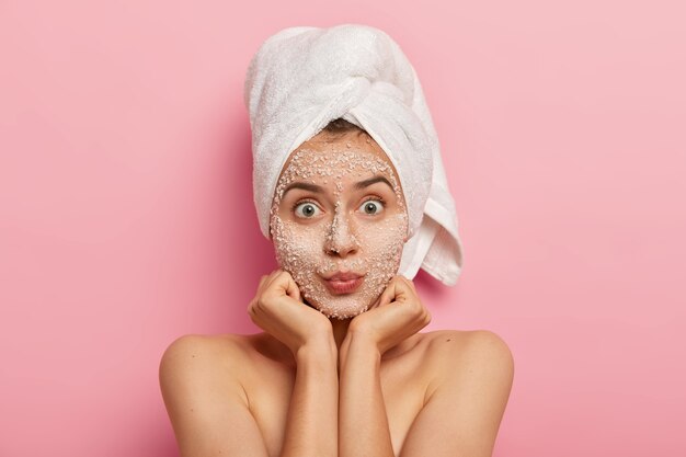 Disparo horizontal de una mujer caucásica sorprendida que mantiene las palmas de las manos debajo de la barbilla, mira con ojos abiertos, aplica mascarilla, evita problemas con la piel, se para sin camisa contra la pared rosada. Concepto de belleza