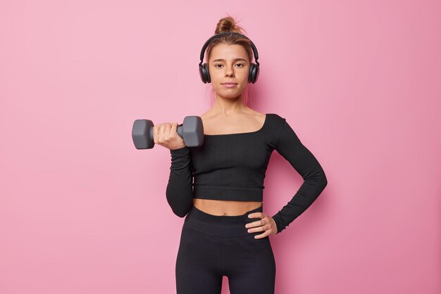 Disparo horizontal de mujer deportiva motivada vestida con top negro y legings levanta mancuernas músculos de trenes interiores escucha música a través de auriculares aislados sobre fondo rosa hace ejercicios deportivos