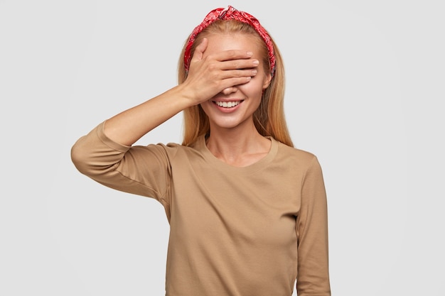 Disparo horizontal de mujer encantadora alegre alegre con expresión feliz, cubre los ojos con la mano, anticipa sorpresa