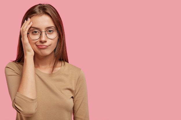 Disparo horizontal de mujer encantadora que ha aburrido la expresión facial disgustada, parece descontento, usa gafas redondas y un suéter informal, aislado sobre una pared rosa con espacio de copia en blanco a un lado