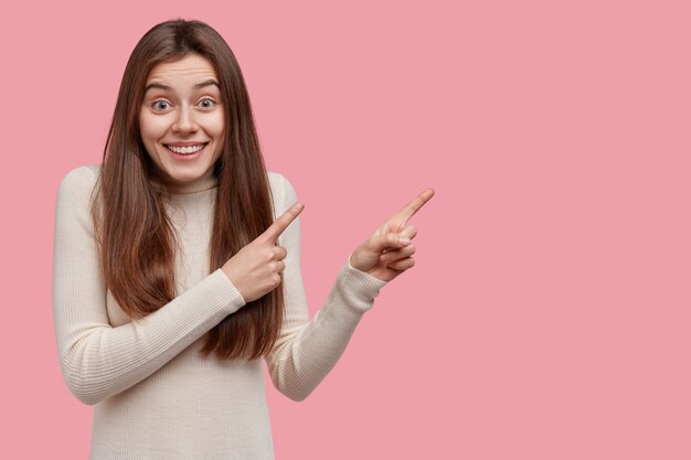 Disparo horizontal de una mujer europea alegre y complacida muestra a los clientes un producto genial, apunta a un lado en la pantalla en blanco, viste ropa casual