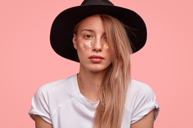 Disparo horizontal de una mujer europea de aspecto agradable que tiene destellos en las mejillas, viste un sombrero negro y una camiseta blanca informal, se ve con confianza, muestra su belleza, aislada sobre una pared rosa