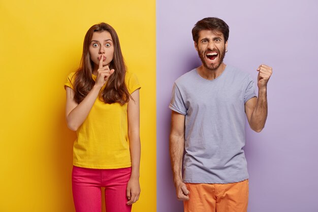 Disparo horizontal de una mujer joven y un hombre juntos en ropa casual, expresan diferentes sentimientos y emociones.La mujer muestra un gesto de silencio con una expresión de sorpresa.Un chico indignado levanta el puño con ira.