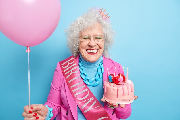Disparo horizontal de mujer mayor complacida sonríe con dientes tiene una apariencia bien cuidada celebra el cumpleaños número 102 disfruta de una compañía alegre se ve hermosa en la vejez sostiene un pastel dulce y un globo de helio inflado