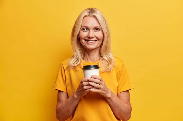 Disparo horizontal de mujer de mediana edad bastante arrugada sostiene una taza de café desechable disfruta del tiempo libre y mira felizmente a la cámara vestida con una camiseta casual