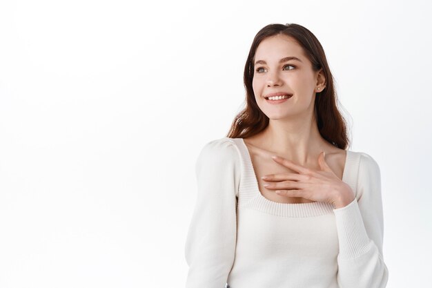 Disparo horizontal de una mujer muy sonriente con una sonrisa encantadora, toca el corazón, se mueve para ser elogiada por alguien, viste ropa casual, se encuentra en el interior de la pared blanca