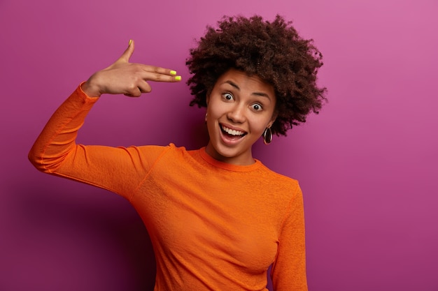 Disparo horizontal de mujer rizada positiva dispara en las sienes, hace gestos de suicidio, hace tonterías, hace pistola de dedo, se ríe alegremente, viste un jersey naranja, aislado en una pared púrpura