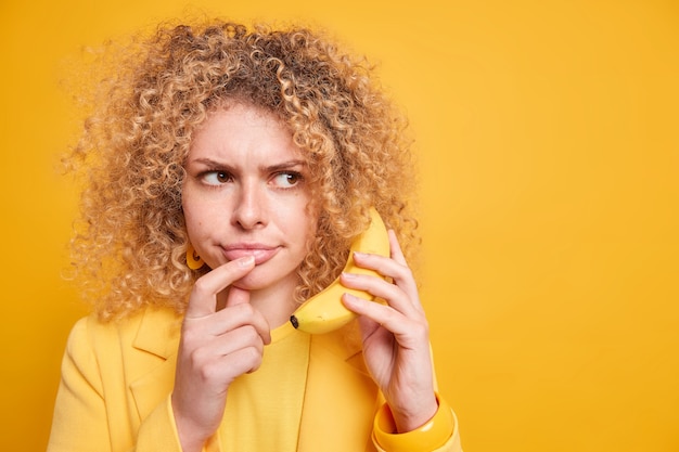 Disparo horizontal de mujer seria disgustada concentrada a un lado sostiene plátano cerca de la oreja finge tener una conversación telefónica discute cuestiones importantes vestidas con ropa formal espacio en blanco en la pared amarilla