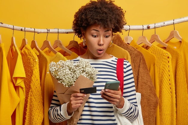 Foto gratuita disparo horizontal de mujer sorprendida rizada usa un teléfono celular moderno, verifica el saldo, tiene tarjeta de crédito, hace compras en línea