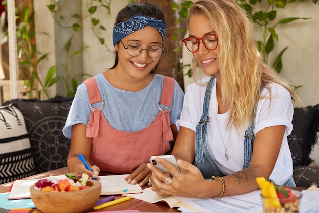 Disparo horizontal de mujeres felices discuten blogs divertidos en Internet, usan el teléfono móvil