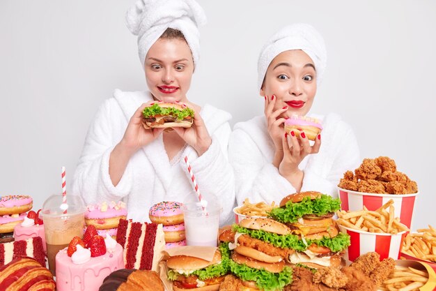 Disparo horizontal de mujeres felices que disfrutan de la fiesta doméstica mantenga la hamburguesa y el donut efford para comer comida chatarra