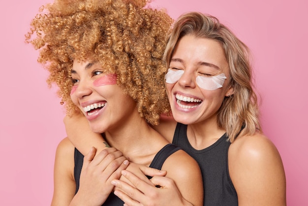 Disparo horizontal de mujeres jóvenes alegres que se abrazan y se divierten aplicar parches de hidrogel de belleza debajo de los ojos cuidar la delicada piel sonreír ampliamente usar camisetas negras aisladas sobre fondo rosa