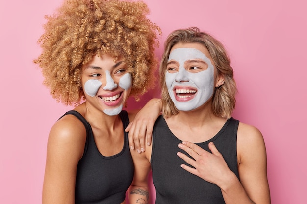 Disparo horizontal de mujeres sinceras felices que disfrutan pasar tiempo juntas, se miran unas a otras, aplican máscaras de arcilla en las caras, se encuentran muy aisladas sobre fondo rosa Concepto de belleza y cuidado de la piel