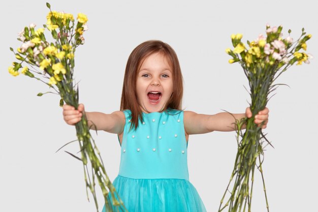 Disparo horizontal de niño pequeño contento sostiene dos ramos de flores, abre la boca abierta, exclama con alegría, viste un vestido azul, aislado sobre la pared blanca.