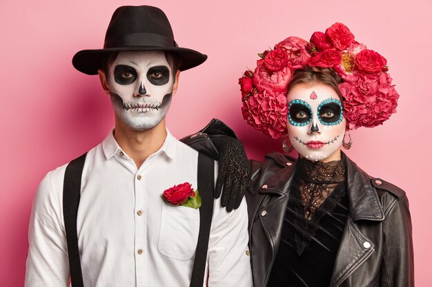 Disparo horizontal de una pareja espeluznante vestida para el Día de Muertos en México, usar máscaras de calaveras, estar uno al lado del otro, celebrar Halloween juntos, aislado sobre fondo rosa