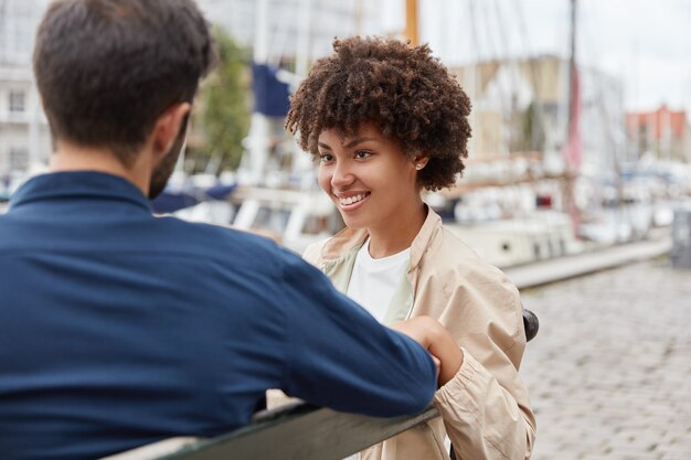 Disparo horizontal de una pareja romántica en el amor sentarse en un banco contra el fondo del puerto marítimo, tener una charla encantadora