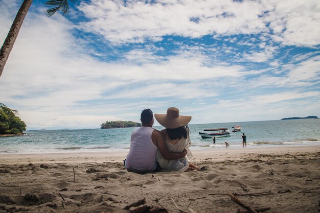 Disparo horizontal de una pareja sentada en una playa hacia las tranquilas aguas azules bajo el hermoso cielo