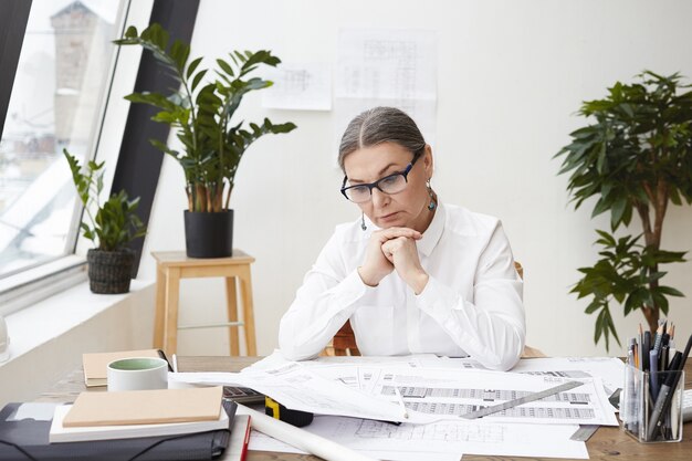 Disparo horizontal de pensativa ingeniera de mediana edad con gafas negras y camisa blanca con las manos juntas debajo de la barbilla, estudiando dibujos y especificaciones en el escritorio frente a ella