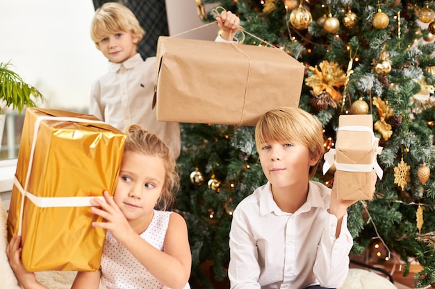Disparo horizontal de tres lindos hermanos sentados en el árbol decorado de Año Nuevo sosteniendo cajas con regalos de Navidad, impacientes, con miradas curiosas. Infancia feliz, alegría y fiesta.