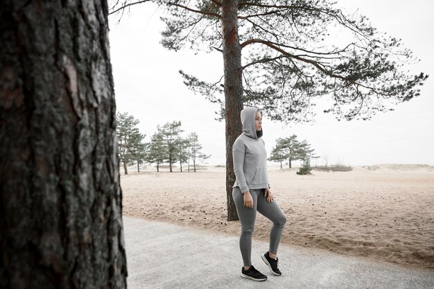 Disparo de longitud completa de una corredora europea de moda fresca que calienta el cuerpo antes del entrenamiento cardiovascular al aire libre, con elegantes zapatillas de deporte, mallas y sudadera con capucha, de pie en un sendero pavimentado en el bosque o el parque