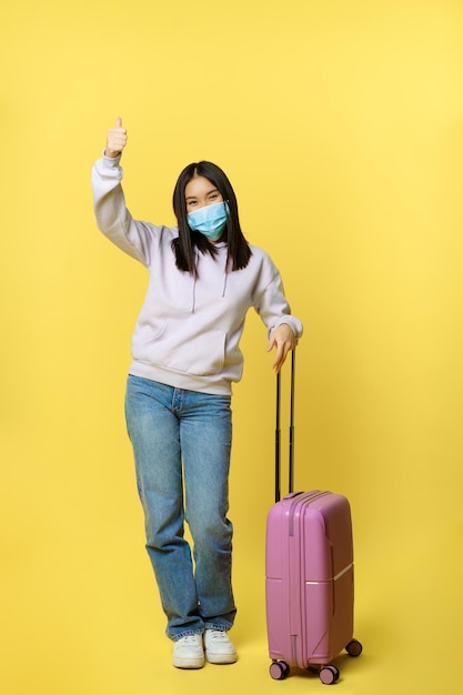 Disparo de longitud completa de una entusiasta chica coreana disfrutando de las vacaciones, posando con maleta, con máscara médica facial, viajando al extranjero durante la pandemia de covid, fondo amarillo.