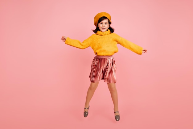 Disparo de longitud completa de linda niña en boina amarilla Foto de estudio de niña preadolescente emocionada saltando sobre fondo rosa