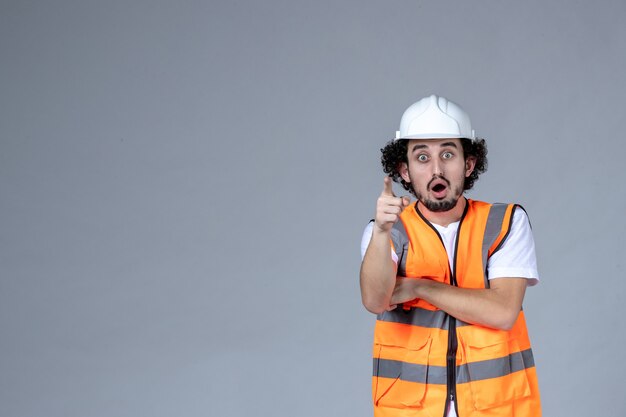 Disparo de medio cuerpo del arquitecto masculino sorprendido en chaleco de advertencia con casco de seguridad y cruzando el brazo sobre la pared gris de la ola