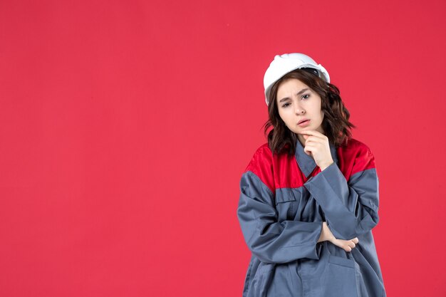 Disparo de medio cuerpo de constructor femenino preguntándose en uniforme con casco y concentrado en algo sobre fondo rojo aislado