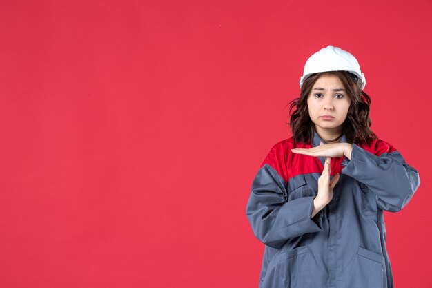 Disparo de medio cuerpo de constructora infeliz en uniforme con casco haciendo gesto de parada sobre fondo rojo aislado