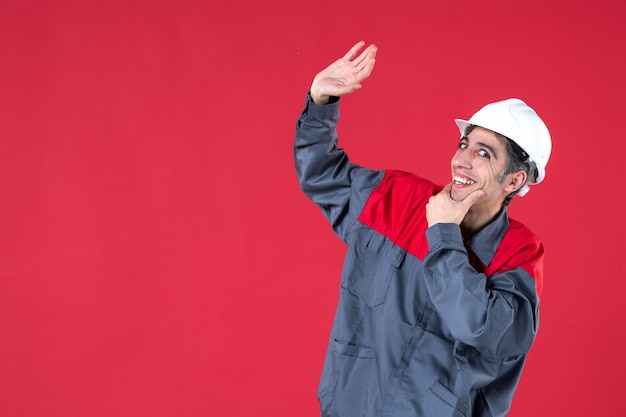 Disparo de medio cuerpo del sonriente joven arquitecto en uniforme con casco y levantando una de sus manos en la pared roja aislada