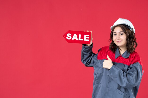 Foto gratuita disparo de medio cuerpo de trabajadora sonriente en uniforme con casco y señalando el icono de venta haciendo gesto ok sobre fondo rojo aislado