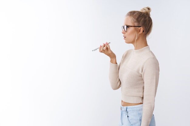 Disparo de perfil de una joven mujer rubia elegante con gafas sosteniendo un teléfono inteligente cerca de la boca que habla altavoz grabando un mensaje de voz sobre fondo blanco con una mirada centrada de aspecto serio y mandona